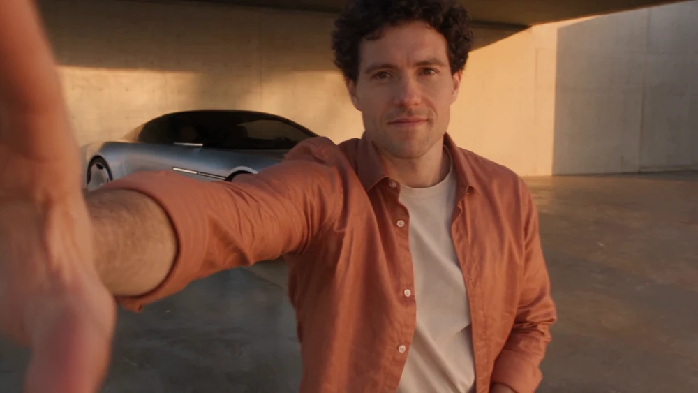 Presenter in a rust shirt reaching away from the lens in front of a fictional silver sports coupe.
