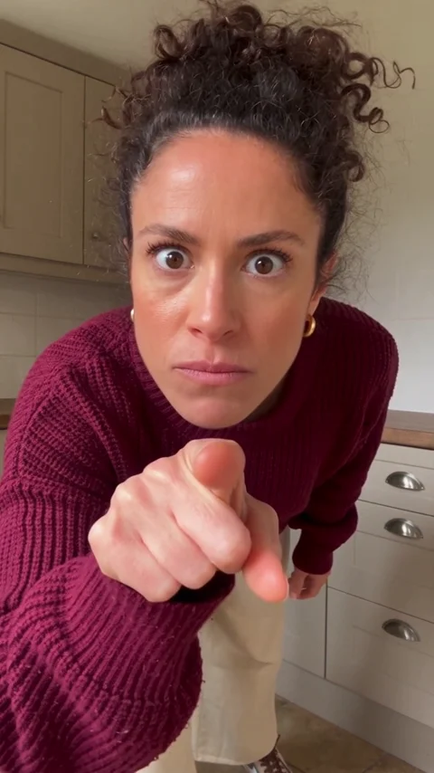 A fictional woman in a burgundy sweater leans close to the phone and points at the lens, with a skeptical expression.