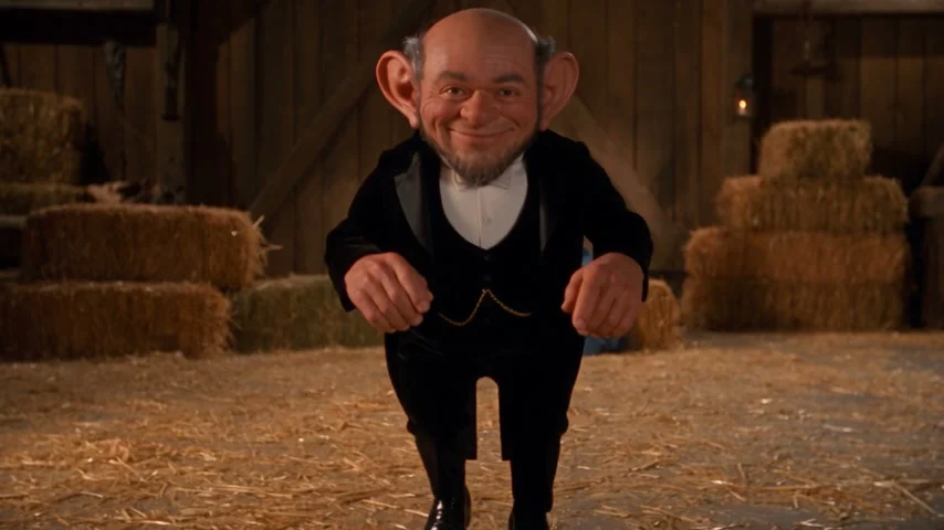 A fictional adult gnome in a black tuxedo tiptoeing in a warm straw-filled barn while an adult woman watches from a hay bale